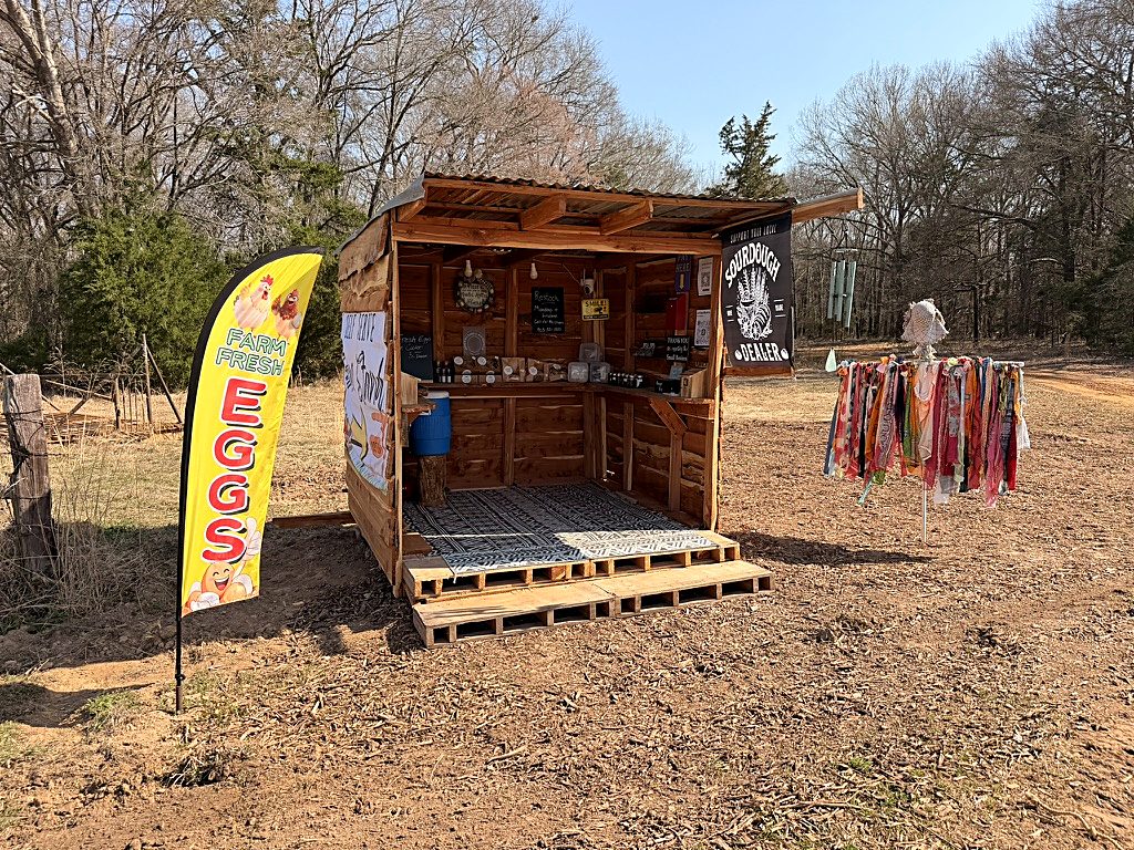 The Honeysuckles & Sourdough wooden farm stand with an EGGS flag and Sourdough Dealer sign in Ben Wheeler, Texas